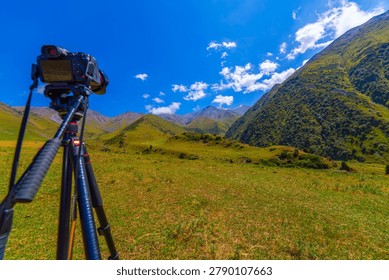 Digital camera mounted on tripod focuses on breathtaking mountain range. Lush green hills and snow-capped peaks under bright blue sky create an inspiring outdoor scene.
