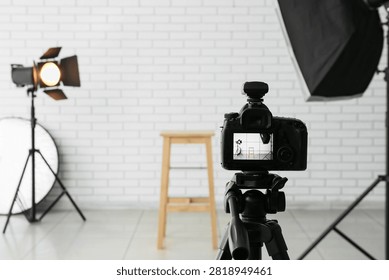 Digital camera on tripod with studio lighting equipment and white brick wall background for photography production, the camera screen displays the scene with a wooden stool and lighting setup