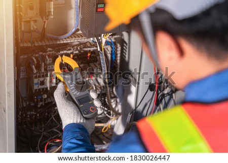 Digital clamp meter in hands of electrician close-up against background of electrical wires and relays. Adjustment of scheme of automation and control of electrical equipment
