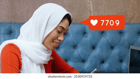 Digital composite of biracial woman with hijab typing on her phone while sitting on a lounge table with a laptop and a cup of coffee. Next to her image of orange message bubble with a heart icon and - Powered by Shutterstock