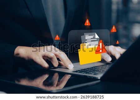 Digital data security breach alert: Close-up of hands using a laptop with a glowing orange warning icon on a file folder, surrounded by red exclamation marks.