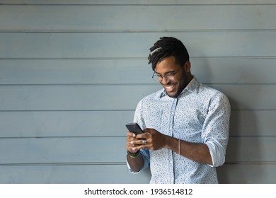 Digital Generation. Confident African Hipster Guy Use Smartphone Chat At Social Network In Good Mood. Smiling Young Black Male Stand On Grey Wall Background Enjoy Surfing Internet On Cell. Copy Space