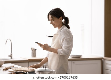 Digital help for home baking. Smiling young woman housewife in kitchen apron stand by countertop with flattened dough for pizza pie watch culinary video guide follow online recipe using mobile phone