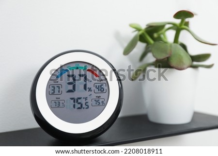 Digital hygrometer with thermometer and green plant on shelf, closeup