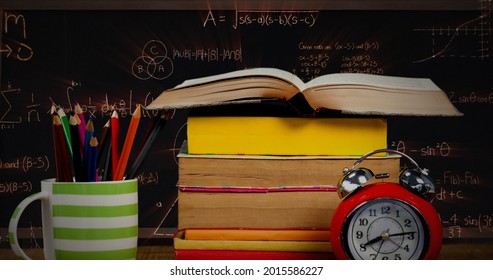 Digital image of a  table with a pile of books, a cup with colorful pens, and a red alarm clock while mathematical equations move in the dark background