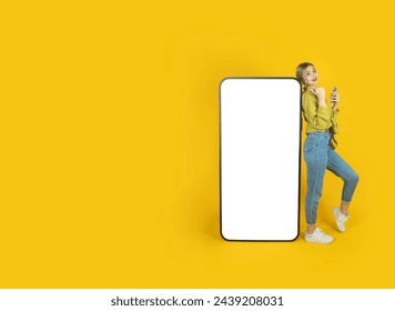Digital mobile application advertisement concept idea image. Full body young woman 20s leaning big empty screen cell phone direct thumb display touchscreen mock up. Yellow studio background copy space