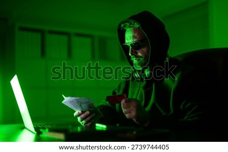 Digital money thief. Hacker programmer in hoodie at office laptop, neon. Hacker attack. Hacker at night in office count money. Cyber attack. Cyber criminal and stolen financial data. Anonymous hacker