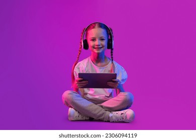 Digital Native, Generation Alpha Concept. Preteen kid happy pretty adorable girl in summer outfit sitting on floor over futuristic background, using wireless headphones and digital tablet, copy space - Powered by Shutterstock