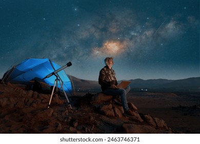 digital nomad man sitting alone with a laptop in the desert at night next to a telescope watching the starry night  - Powered by Shutterstock