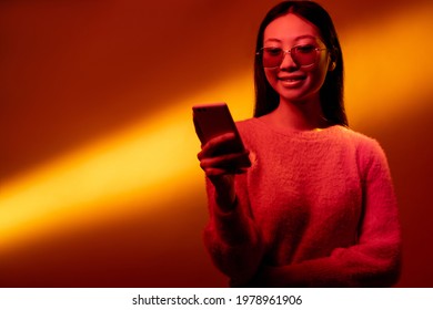 Digital People. Mobile Technology. Color Light Portrait. Online Communication. Neon Pink Smiling Asian Girl Using Phone Isolated On Yellow Golden Gradient Copy Space Background.