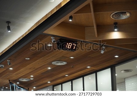 Digital queue number display showing number 187 mounted on a modern wooden ceiling interior with spotlights, CCTV cameras, and ventilation system.