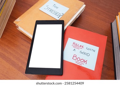 Digital reading device with blank screen on books with sticky notes with the texts ¨too stressed?¨ and ¨Relax with a good book¨. Black e-reader on a wooden table next to some books
