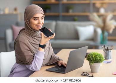 Digital Speaker Application. Portrait of young muslim woman in hijab sitting at table, talking on mobile phone, using laptop and virtual voice assistant for online search or to record reminder - Powered by Shutterstock