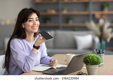 Digital Speaker Application. Portrait of young smiling woman sitting at table, talking on mobile phone, using laptop and virtual voice assistant for online search, to record reminder, dictate message - Powered by Shutterstock