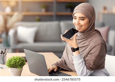 Digital Speaker Application. Portrait of young muslim woman in hijab sitting at table, talking on mobile phone, using laptop and virtual voice assistant for online search or to record reminder - Powered by Shutterstock