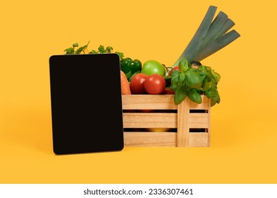 Digital Tablet With Blank Screen Standing Near Wooden Box With Fresh Vegetables And Fruits Over Yellow Studio Background. Mockup For Agriculture Website Advertisement Or Online Grocery Shopping
