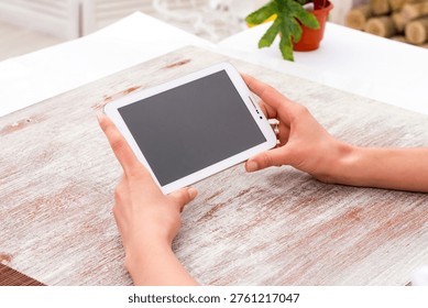 Digital tablet computer with blank screen held in woman hands over table, modern device mockup for apps, UI design, and digital work concepts.
