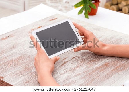 Digital tablet computer with blank screen held in woman hands over table, modern device mockup for apps, UI design, and digital work concepts.