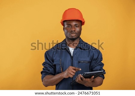 Digital tablet in hand, construction worker. Handsome black man is in the studio against yellow background.