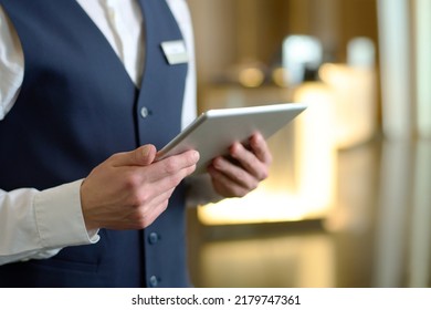 Digital tablet in hands of young male receptionist of luxurious modern hotel standing in lounge by entrance and meeting new guests - Shutterstock ID 2179747361