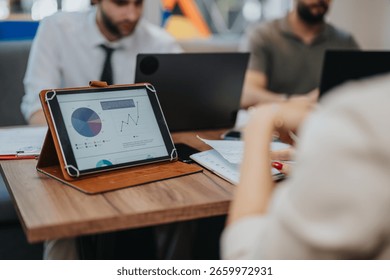 A digital tablet showcasing data graphs sits on a table, surrounded by coworkers engaged in collaborative work, emphasizing teamwork and analytics in a business setting.