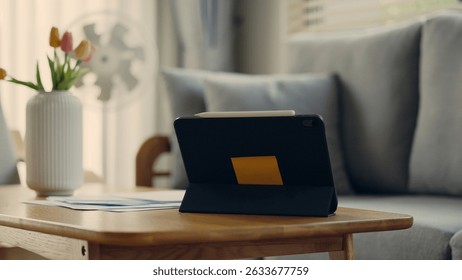Digital tablet with stylus placed on wooden coffee table in cozy home setting. Modern lifestyle, remote work or creative space with flowers and soft daylight.