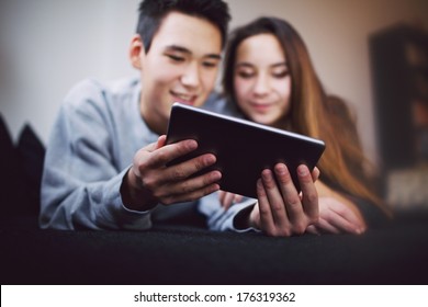 Digital Tablet In Young Man's Hand Lying With His Girlfriend On Couch. Teenage Couple Looking At Tablet PC. Mixed Race Man And Woman Reading Email On Touch Screen Computer At Home.