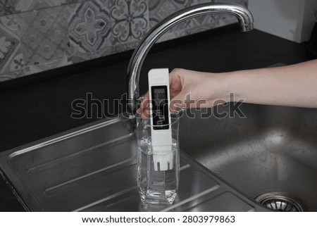Digital TDS meter measuring tap water collected in a drinking glass beside a kitchen sink. Concept of water quality testing, tap water, dissolved solids, impurities, consumer safety.