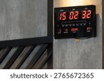 Digital wall clock. Red LED numbers contrast against dark casing. Clock mounted on textured gray wall beside black railing