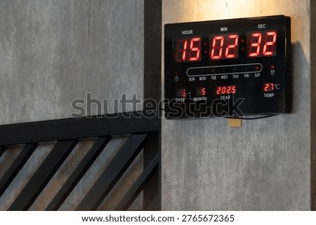 Digital wall clock. Red LED numbers contrast against dark casing. Clock mounted on textured gray wall beside black railing