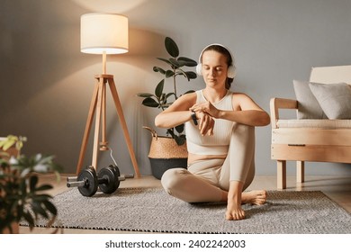 Digital wellbeing at home. Smart bracelet in sport. Sporty smartwatch features. Sportswoman's tracker. Sporty beautiful woman in headphones checking her workout time at home - Powered by Shutterstock