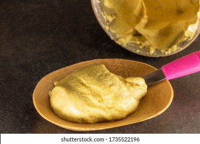 Dijon Mustard In A Wooden Ramekin With A Pink Tea Spoon And A Large Jar Of Mustard In The Background