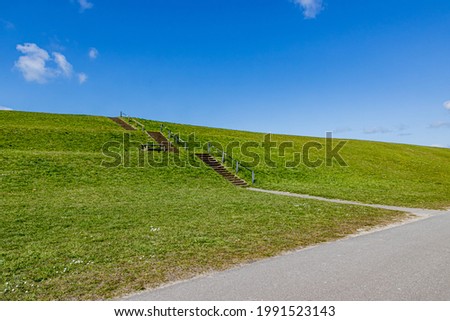 Dike on the beach of Camperduin with green grass and concrete stairs with a metal railing, sunny spring day with a blue sky and some white clouds in North Holland in the Netherlands