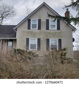 A dilapidated old farmhouse with weathered off-white siding and brown shutters stands in an overgrown yard with tall grass and unkempt shrubbery against a gray-blue sky. - Powered by Shutterstock