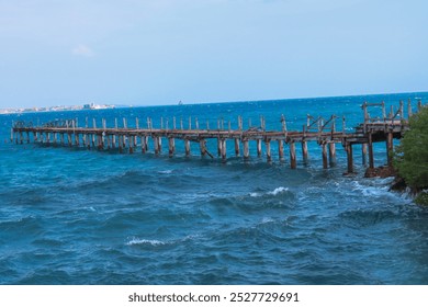 A dilapidated wooden pier extends into the ocean near Prison Island, Zanzibar. The waves crash against the structure, with a distant view of the mainland and ships visible on the horizon. - Powered by Shutterstock