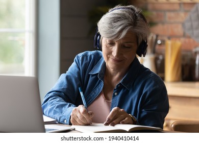 Diligent aged student. Focused elderly hispanic woman in headphones watch webinar on laptop screen take notes to paper notebook. Mature old lady in headset study online take part at education training