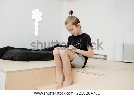 Diligent boy is sitting on a step of a stair to elevated floor, writing something on his knees.