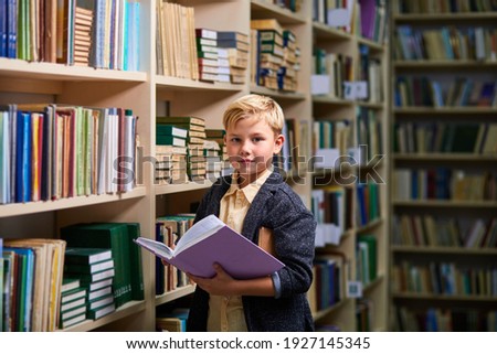 diligent child boy with book between bookshelves in campus library, he is looking at camera. learning, brain, education concept