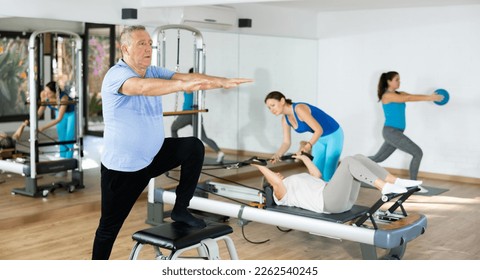 Diligent elderly man practicing pilates on pedal fitness chair in sports hall during pilates classes. Persons doing pilates with trainer - Powered by Shutterstock