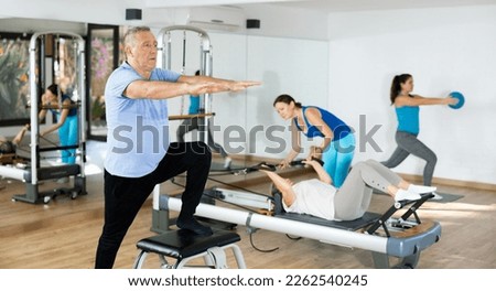 Diligent elderly man practicing pilates on pedal fitness chair in sports hall during pilates classes. Persons doing pilates with trainer