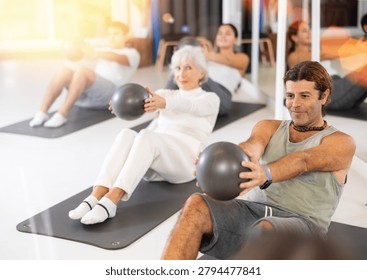 Diligent middle-aged man practicing pilates with ball in training area during pilates classes