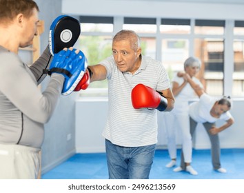 Diligent old man training boxing kicks on punch mitts held by instructor in sports hall - Powered by Shutterstock