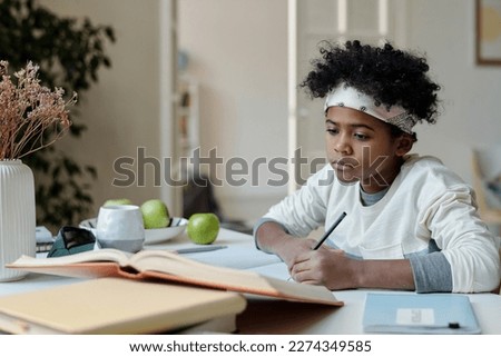 Diligent schoolboy looking through article in encyclopaedia and making notes in copybook while sitting by desk and preparing homework