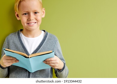 Diligent Schoolboy Reading A Book Over Green Background, Isolated, Copy Space. Smart And Intelligent Blond Child In Gray School Wear Stand Smiling At Camera