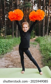 A Diligent, Smiling Girl, A Child Cheerleader In A Black Suit Is Dancing In The Woods In Nature With Big Orange Pom-poms In Her Hands. Sports Training For Cheerleading.