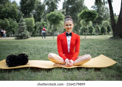 A Diligent, Smiling Girl, A Child Cheerleader In A Red Suit Is Exercising In The Woods In Nature With Big Pom-poms In Her Hands. Sports Training For Cheerleading.