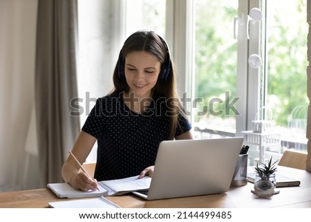 Diligent student in headphones makes task sit at desk with laptop, e-learn english, take part in video call with online tutor, listen audio course looks happy enjoy study. Learning using tech concept