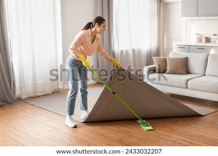 Diligent woman in casual attire meticulously lifts corner of her area rug to ensure thorough clean with bright green mop in sunlit living room, full length