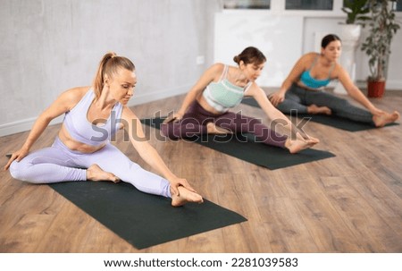 Diligent women practicing hand to toe pose of yoga on black mat in light fitness room with house plants