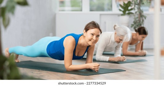 Diligent women practicing plank pose of yoga on black mat in light fitness room with house plants - Powered by Shutterstock